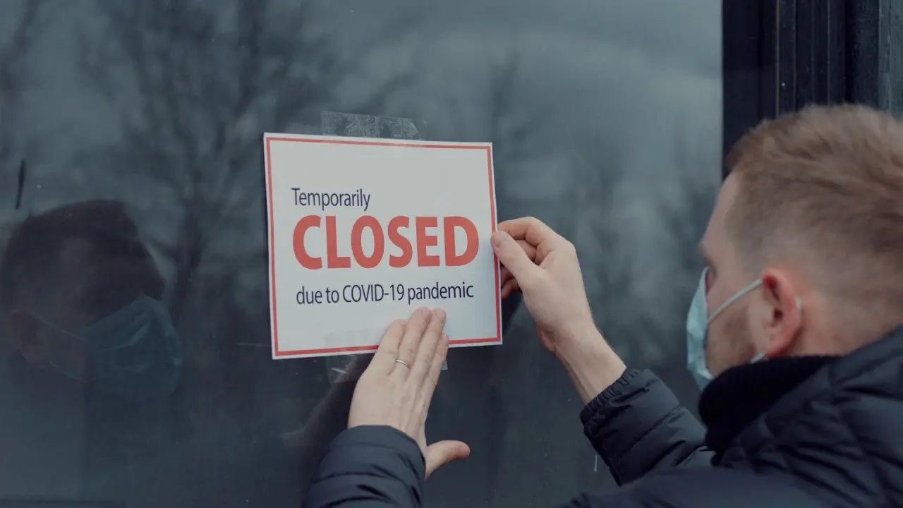 a person wearing a face mask puts up a sign on a door stating, temporarily closed due to COVID-19 pandemic